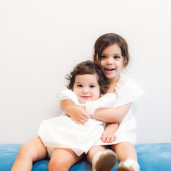 Two young sisters in white dresses hugging and smiling in Dripping Springs, TX